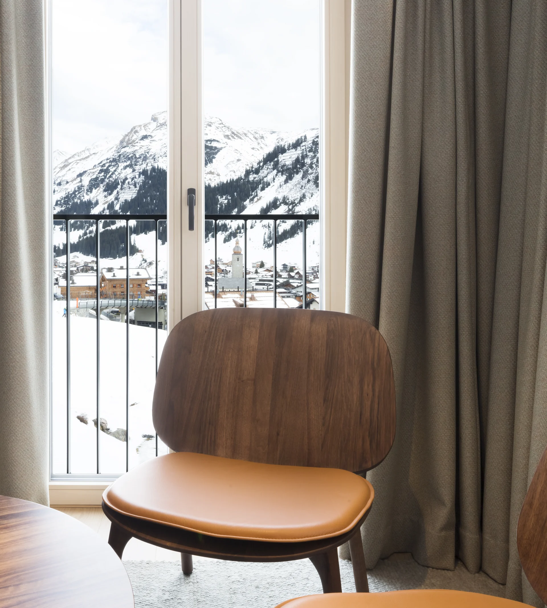 Ihr Hotel in Lech am Arlberg. Das Sunora. Holzstuhl vor Fenster mit Blick auf verschneite Berge und Dorf