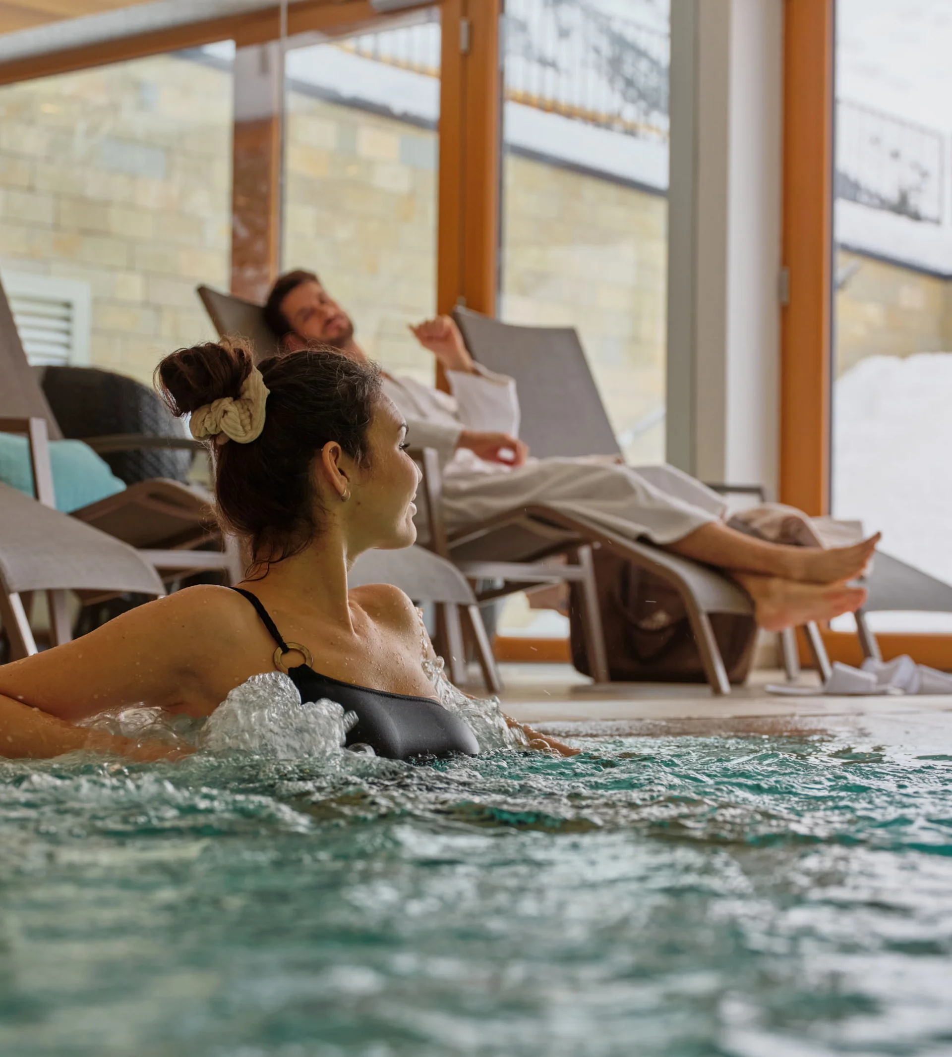 Ihr Hotel in Lech am Arlberg. Das Sunora. Frau entspannt im Whirlpool, Mann liegt auf Liegestuhl im Innenbereich mit großen Fenstern