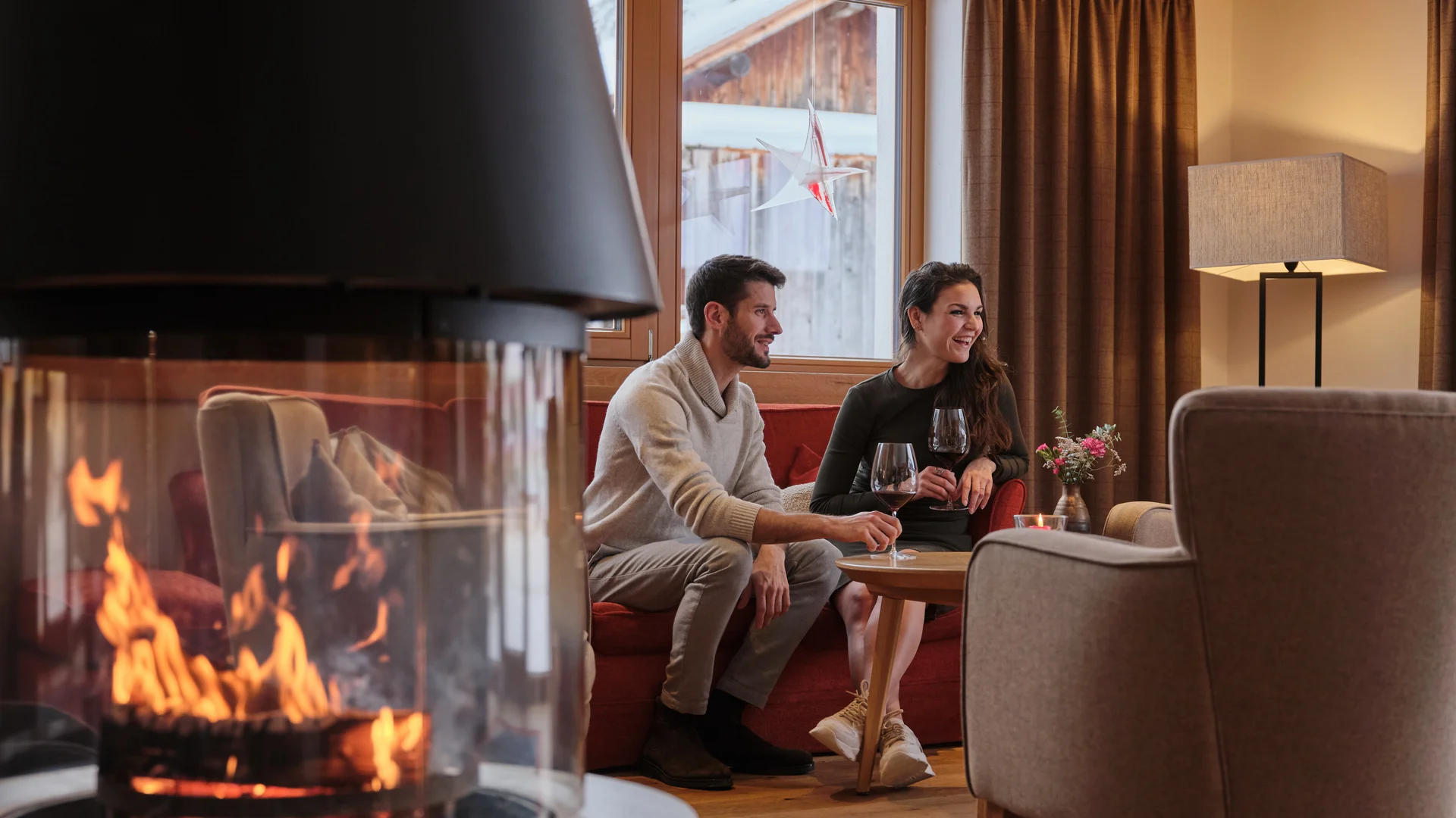 Ihr Hotel in Lech am Arlberg. Das Sunora. Paar sitzt mit Wein vor einem Kamin in einem gemütlichen Wohnzimmer