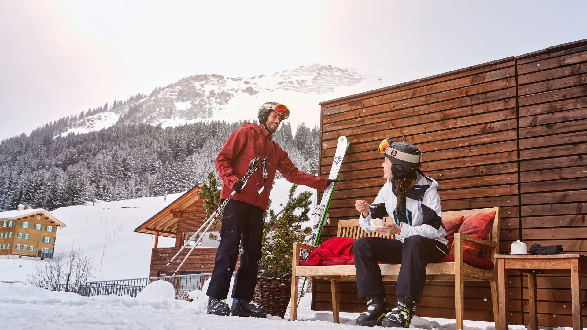 Ihr Hotel in Lech am Arlberg. Das Sunora. Zwei Skifahrer im Winter vor einer Hütte mit schneebedeckten Bergen im Hintergrund