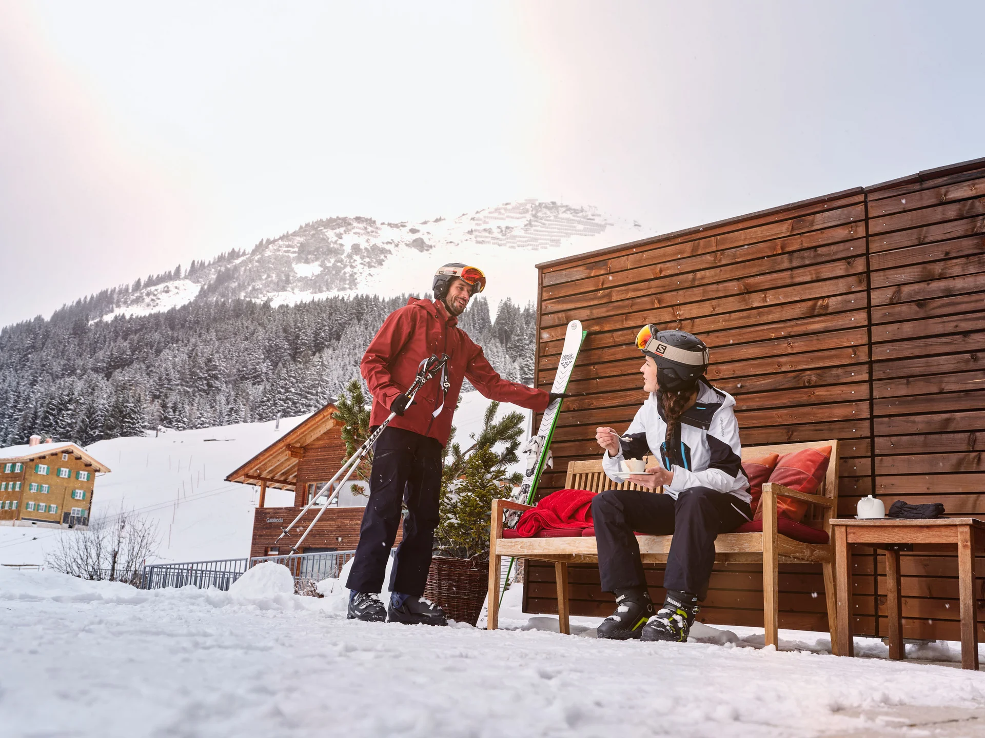 Ihr Hotel in Lech am Arlberg. Das Sunora. Zwei Skifahrer im Winter vor einer Hütte mit schneebedeckten Bergen im Hintergrund