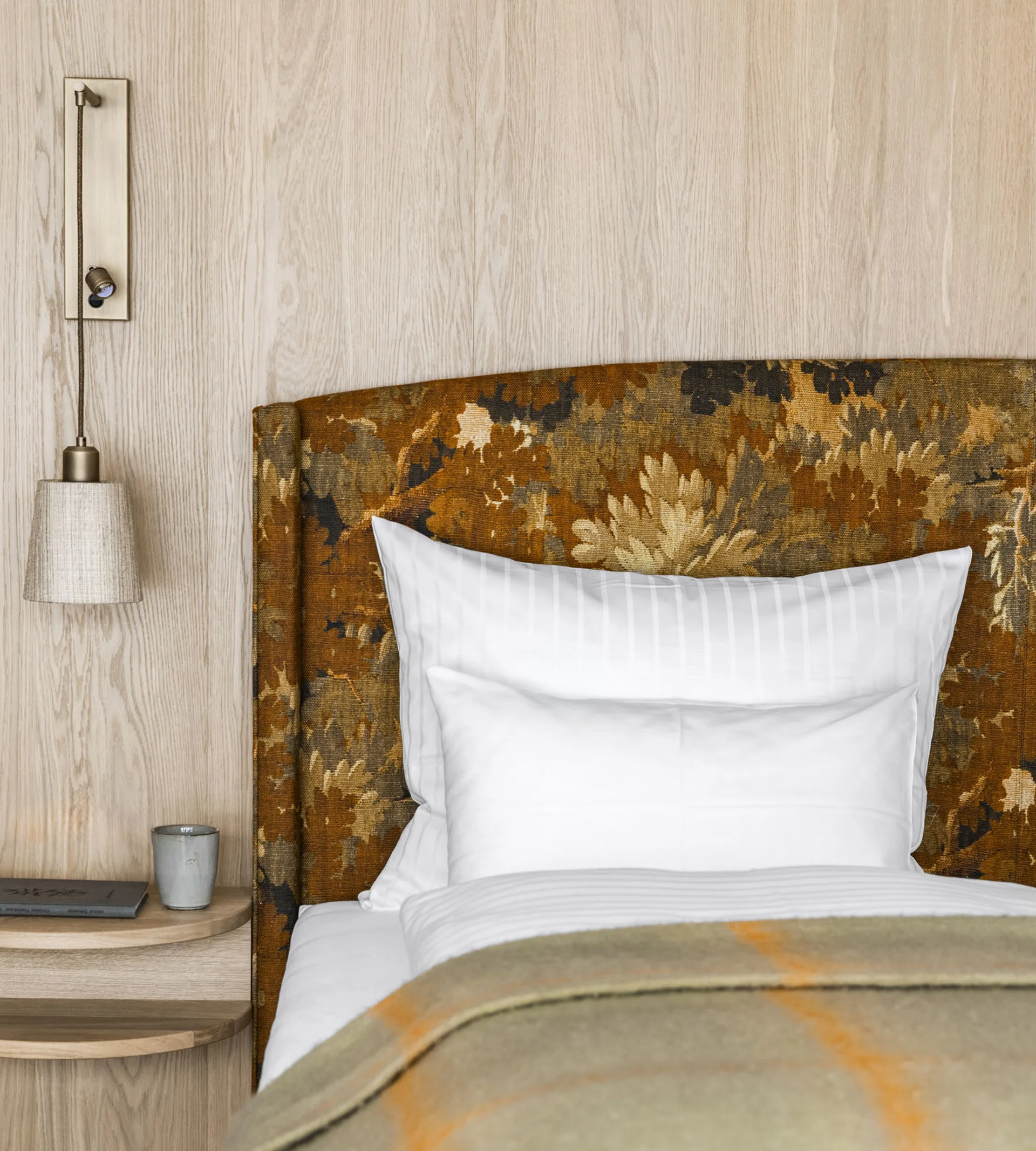 Ihr Hotel in Lech am Arlberg. Das Sunora. Nahaufnahme eines Bettes mit gemustertem Kopfteil und Nachttisch mit Lampe und Buch