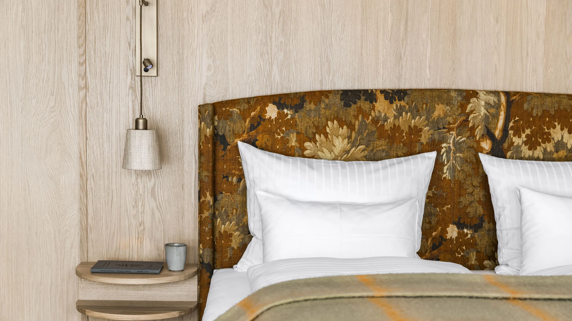 Ihr Hotel in Lech am Arlberg. Das Sunora. Nahaufnahme eines Bettes mit gemustertem Kopfteil und Nachttisch mit Lampe und Buch