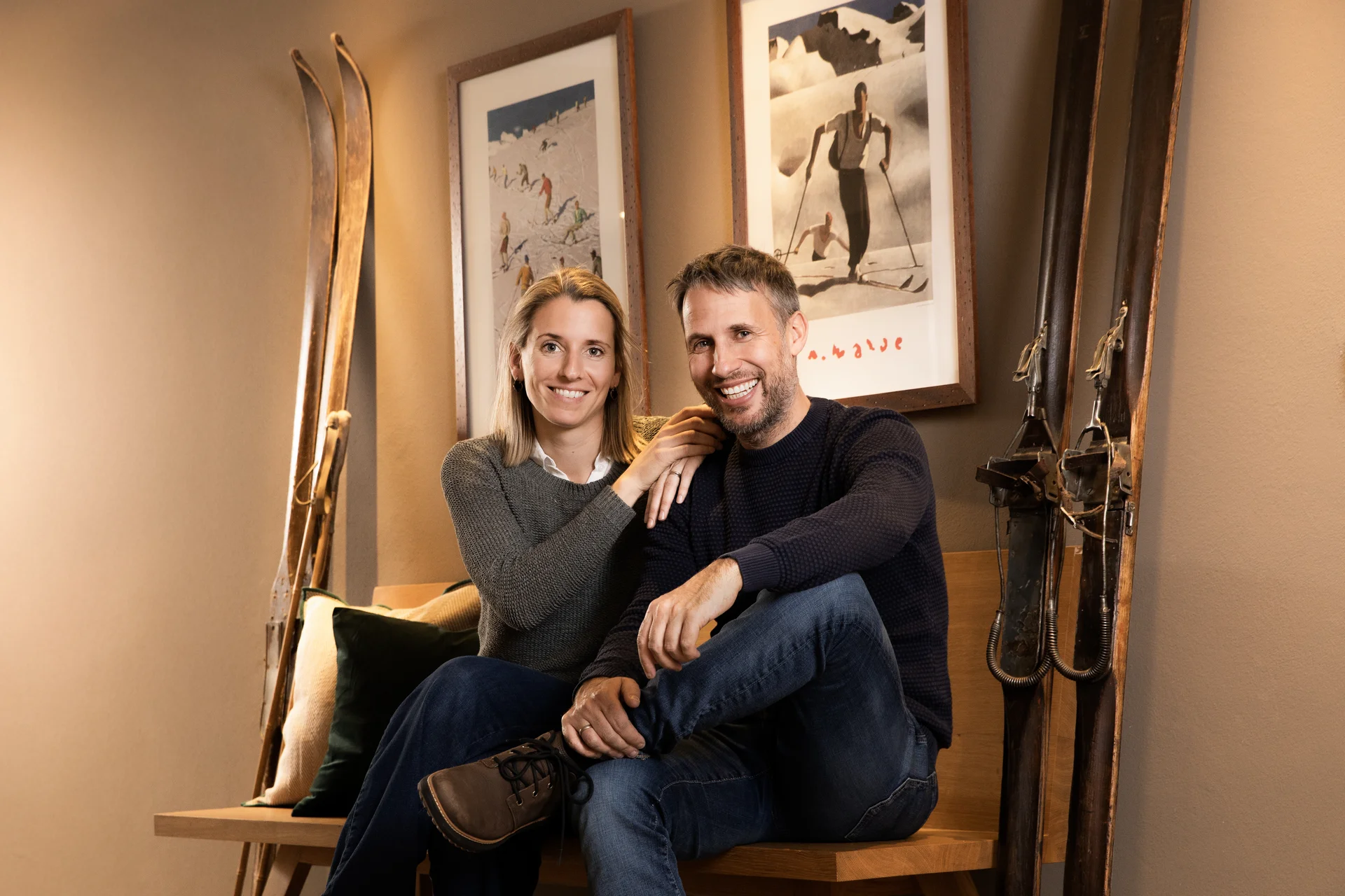 Ihr Hotel in Lech am Arlberg. Das Sunora. Lächelndes Paar sitzt auf einer Holzbank in einem Raum mit Skiern und Wintersportbildern