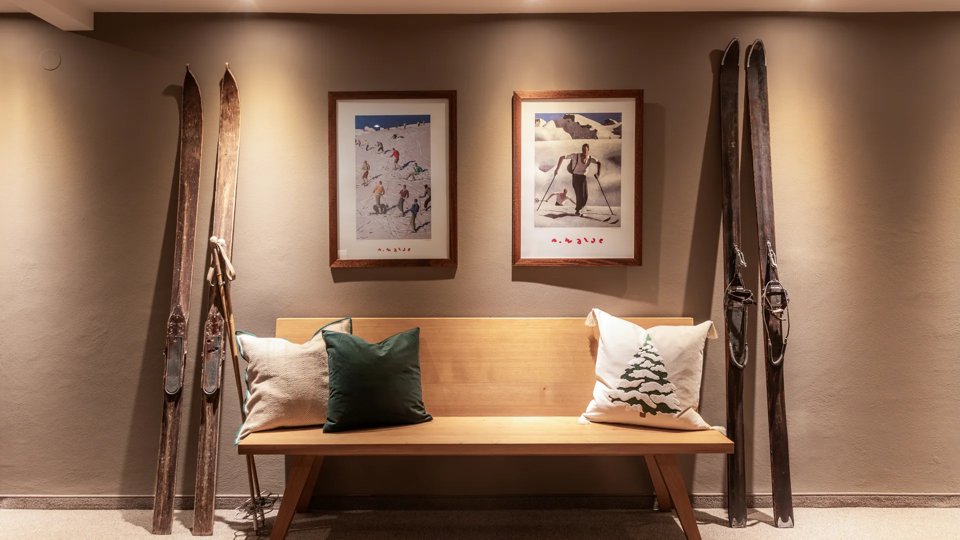 Ihr Hotel in Lech am Arlberg. Das Sunora. Holzbank mit Kissen unter zwei gerahmten Skifahrer-Bildern und antiken Skiern an der Wand