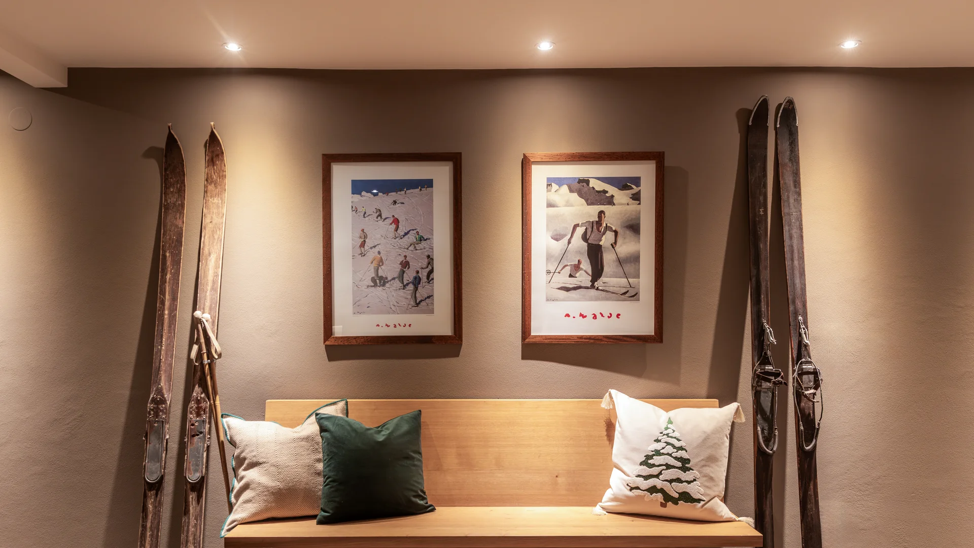Ihr Hotel in Lech am Arlberg. Das Sunora. Holzbank mit Kissen unter zwei gerahmten Skifahrer-Bildern und antiken Skiern an der Wand