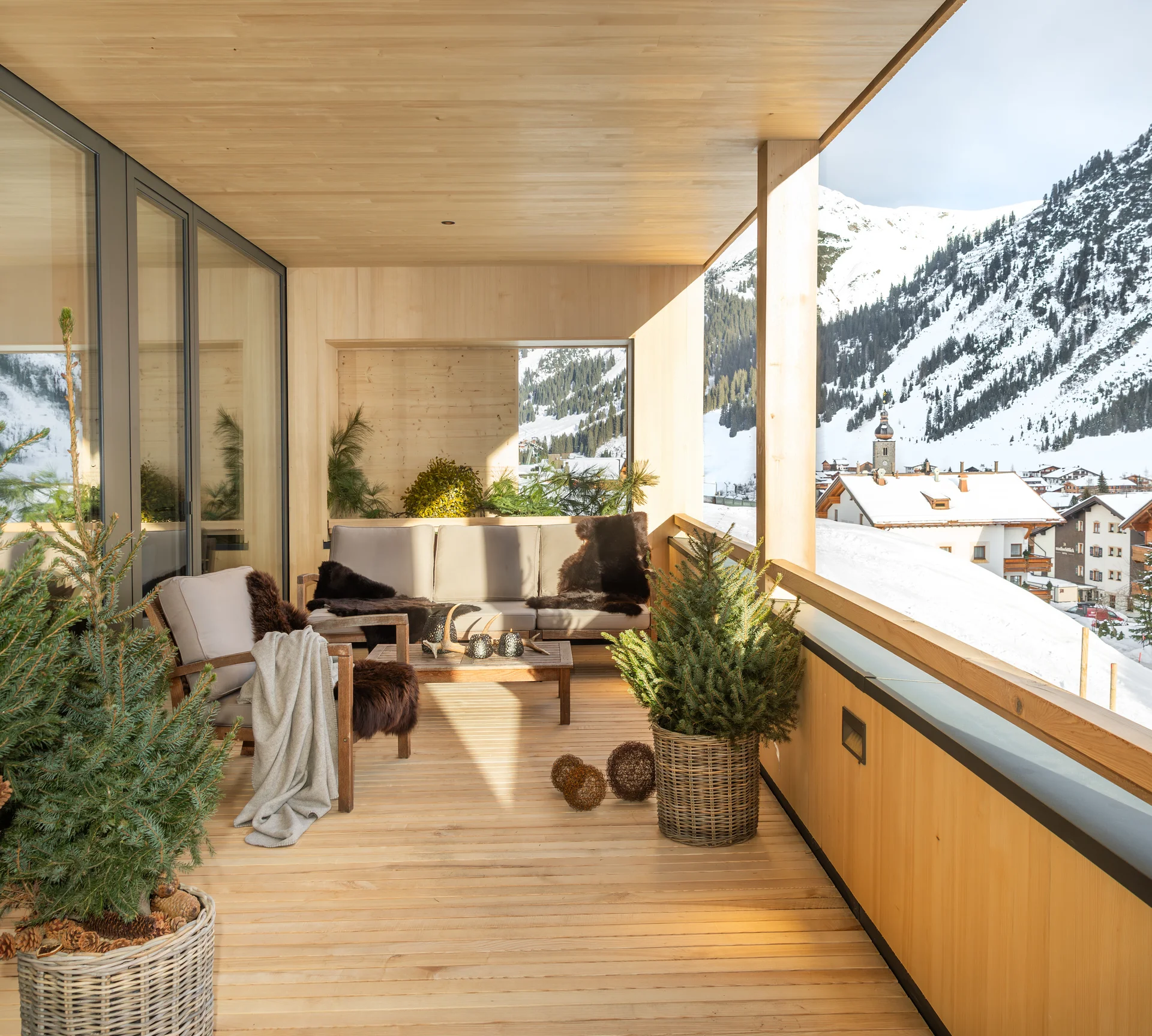 Ihr Hotel in Lech am Arlberg. Das Sunora. Überdachter Holz-Balkon mit Sitzmöbeln und Blick auf verschneite Berge und Dorf