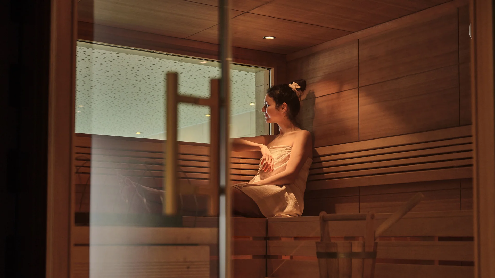 Ihr Hotel in Lech am Arlberg. Das Sunora. Frau entspannt in Sauna, sitzt auf Holzbank mit Handtuch